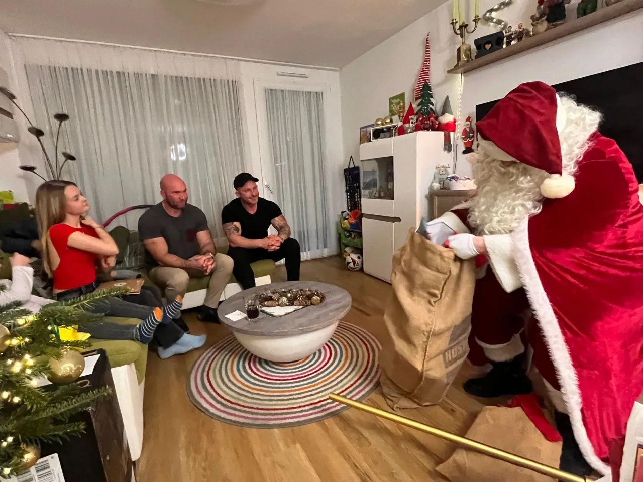 Weihnachtsmann der in seinem Sack nach Geschenken sucht und eine Familie die auf dem Sofa sitzt und gespannt wartet.