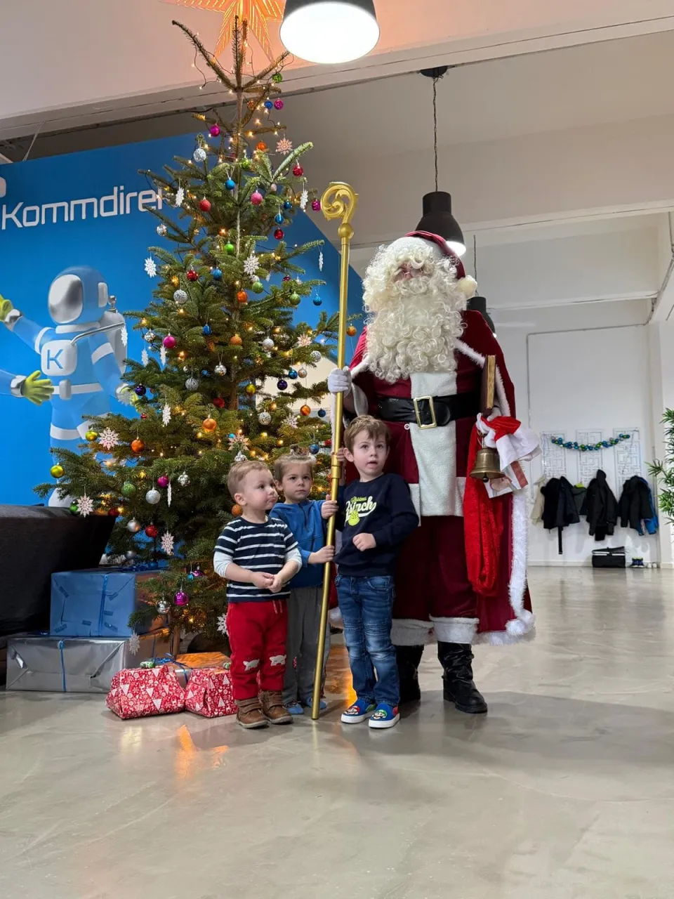 Weihnachtsmann und drei Kinder die vor einem Weihnachtsbaum stehen, der Weihnachtsmann hält seinen Stab und die Kinder helfen ihm dabei.