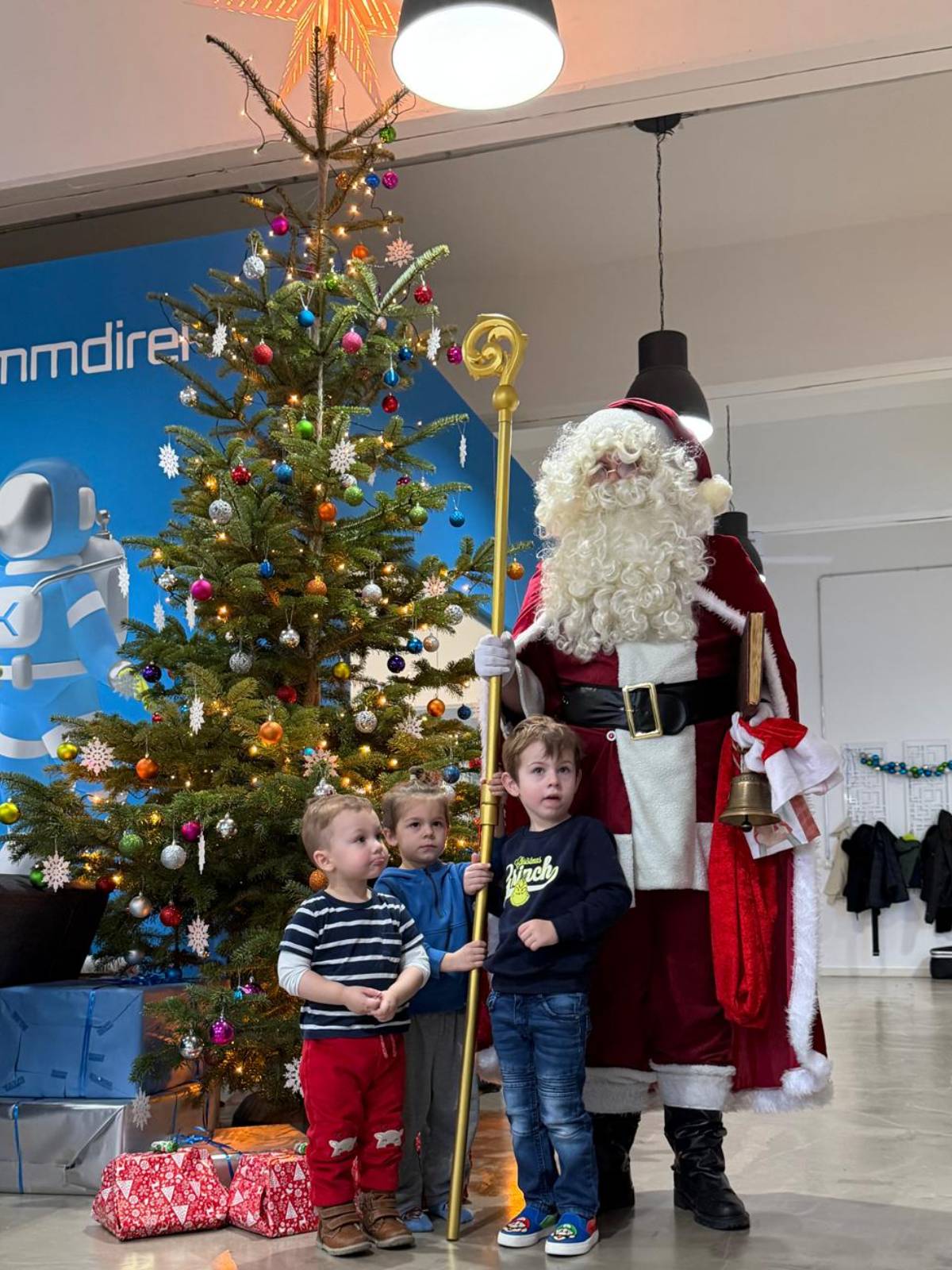 Weihnachtsmann bei einem Gruppenbesuch mit mehreren Kindern Weihnachtsmann steht mit drei Kindern vor einem Weihnachtsbaum, die Kinder helfen ihm seinen Stab zu halten